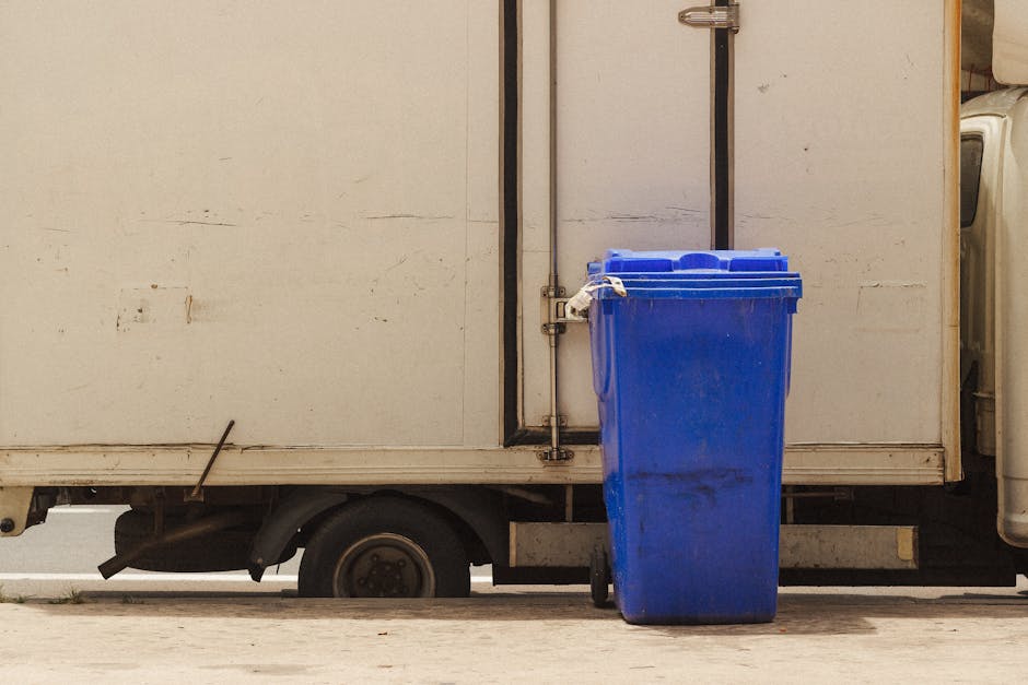 A bright blue recycling bin placed outside a white delivery truck, with the bin's lid closed and slightly dirty marks visible on its surface. The truck appears to be parked on a street or driveway, wi