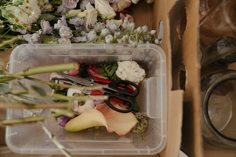 A clear plastic container filled with floral waste including various cut flower stems, petals, and leaves, with a pair of black scissors resting inside. Surrounding the container are loose flowers and