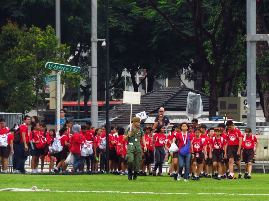 A group of school children in red uniforms and backpacks, accompanied by an adult in military camouflage attire holding a sign, are walking in a line across a grassy field during daytime. The children