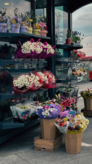 A variety of fresh floral arrangements and bouquets are displayed on dark grey shelving units outside a flower shop, with some wrapped in pastel-colored paper and ribbon, while others are in brown and