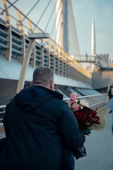 A man dressed in a dark jacket holding a bouquet of fresh flowers in front of a modern bridge structure with steel railings and cables, set against a bright sky. The bouquet features pink and white ro
