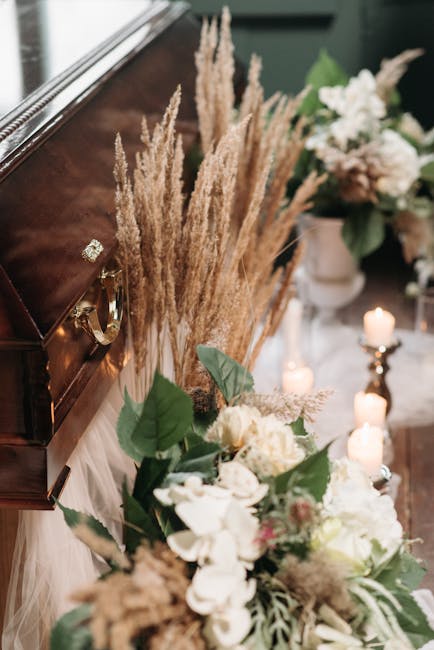 A close-up of a floral arrangement placed on a wooden surface, featuring a bouquet of white and cream-colored flowers including roses and orchids, accented with lush green leaves. Behind the bouquet, 