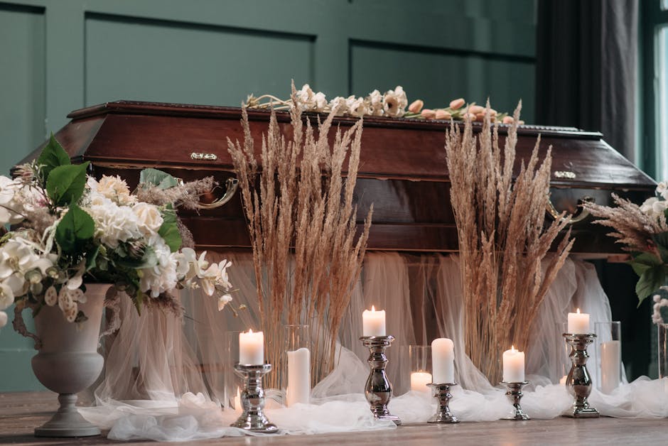 A dark polished wooden casket surrounded by arrangements of white and pale pink flowers, including roses and hydrangeas, placed in elegant white vases. Tall, dried pampas grass and soft pink roses ado
