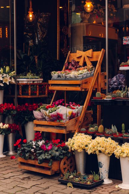 A display of fresh flowers arranged in wooden crates and metal buckets outside a florist shop, with a variety of blooms including red, pink, and white flowers, some wrapped in tissue paper. The arrang