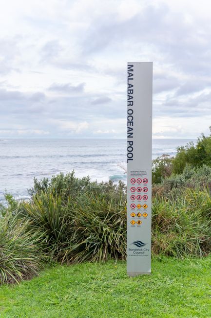 A tall, rectangular signpost reading 'Malabar Ocean Pool' situated on a grassy area near the coast. Behind the sign, the ocean is visible with gentle waves under a cloudy sky. The foreground features