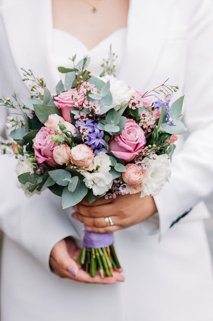 A person dressed in a white outfit holds a rounded bouquet of fresh flowers, including pink roses, white blooms, purple flowers, and sprigs of eucalyptus with silvery-green leaves. The bouquet is wrap