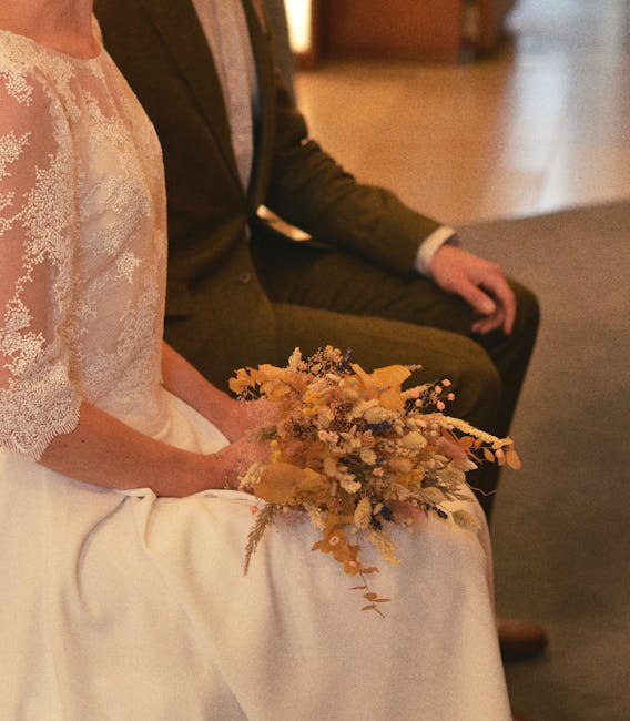 A bride seated in a wedding gown with lace detailing on the bodice, holding a delicate bridal bouquet composed of soft pastel and neutral-toned flowers, including dried and fresh blooms such as roses,