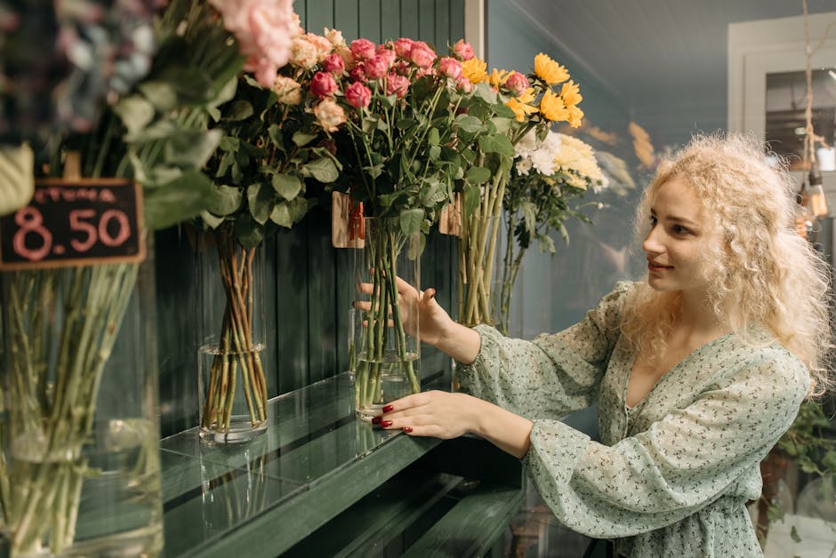 A woman with curly blonde hair and fair skin, dressed in a light, patterned blouse, is arranging a tall, clear glass vase filled with fresh, vibrant pink and yellow flowers on a dark green shelf insid